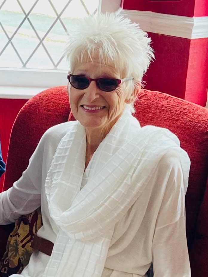 Patricia Harford smiling, seated in a red chair and wearing a white outfit and sunglasses.